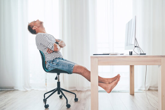 A middle-aged man relaxing on a swivel chair near the table with computer while he watching TV in living room. He stretching barefoot lags and resting. Streaming on worldwide quarantine time concept