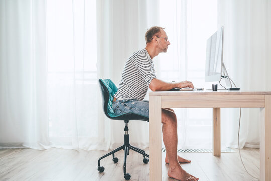 Barefoot Middle-aged writer man on a swivel chair typing down a text using the modern PC keyboard in the home living room. Novelist freelance on worldwide quarantine time concept