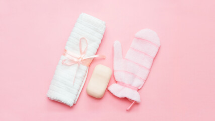 White towel with ribbon, soap and pink exfoliating hydro glove on a delicate pink background. Cosmetology, spa cosmetic products, hygiene, personal care