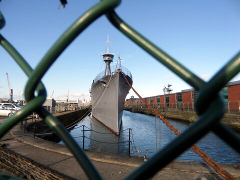 Sehr d&uuml;nnes graues milit&auml;risches Kampfschiff positioniert in der Masche eines gr&uuml;nen Maschendrahtzaunes. Schiffelandschaft Kriegsschiff