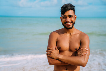 Fototapeta na wymiar Athletic young latin american man on the beach with arms crossed. smiling man looking at camera