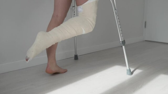 Close-up of women's legs, a woman in plaster up to the hip slowly goes to the door on crutches.