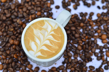 white cup of coffee with an ornament of foam photographed from above placed on a white tablecloth and coffee beans spilled on it