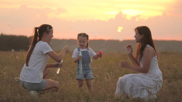 Happy family mom and children play together, blow and catch soap bubbles in field. Mother, little daughters play together in park at sunset. Family, childhood. Soap bubbles in sun, childrens birthday