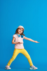 little girl in summer clothes and headphones poses on blue background in full growth and looks to side and points her hand to side. concept summer and spring casual children's clothing.