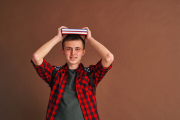 surprised, joyful teenage student in casual clothes with a backpack, holding books on his head, isolated against a brown background. Higher school education, the concept of a university college.