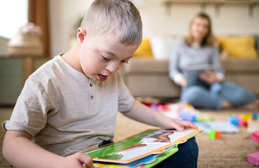 Down syndrome child reading book at home, mother working in home office.