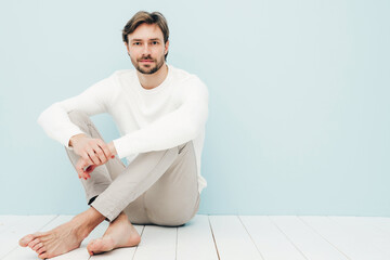 Portrait of handsome smiling hipster lumbersexual businessman model wearing casual white sweater and trousers. Fashion stylish man sitting against light blue wall in studio