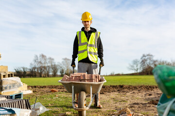 A young adult male builder wearing a high visibility vest and hard hat pushing a wheelbarrow full of bricks while on a building site