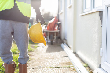 A close up of a unknown builder wearing a high visibility vest and holding a hard hat. Builder,...