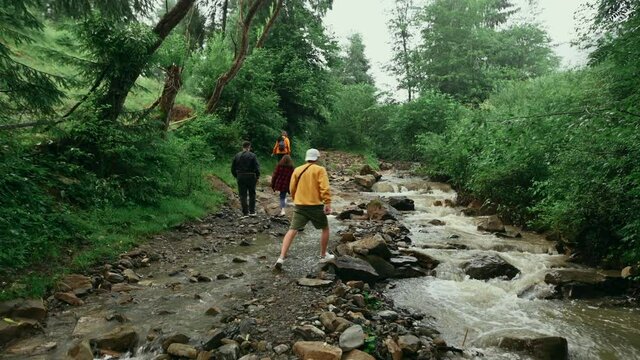 Group of young hikers walking along a forest path in the mountains near a stream. Friends in casual clothes on a hike go through the woods by the river.