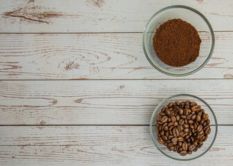 On a light wooden background, there are two bowls with ground coffee and coffee beans. Top view