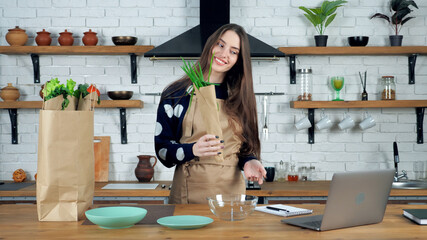 Smiling woman housewife in beige apron shows teacher onions ingredients to webcam study online remote video call conference laptop computer, distance culinary chef cook course lesson in home kitchen