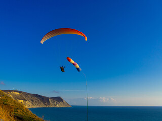 The cone is a wind designator for paragliding in the sky. A tandem paraglider flies over the sea with mountains at sunset.