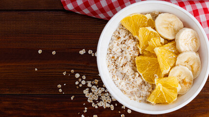 Bowl of oatmeal porridge with banana and orange  on wooden background. Top view, copy space