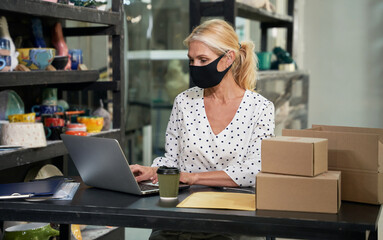 Pandemic. Busy mature woman, small business owner wearing protective mask working on the laptop, receiving orders while sitting at the desk in her craft pottery shop