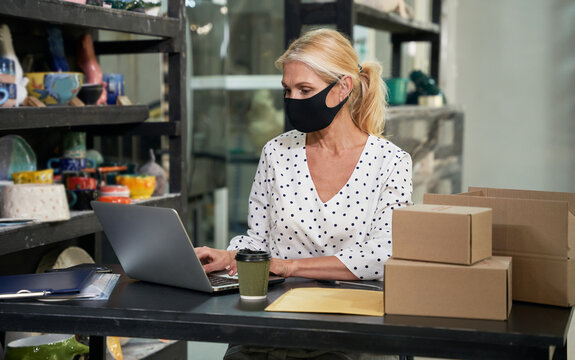 Pandemic. Busy mature woman, small business owner wearing protective mask working on the laptop, receiving orders while sitting at the desk in her craft pottery shop