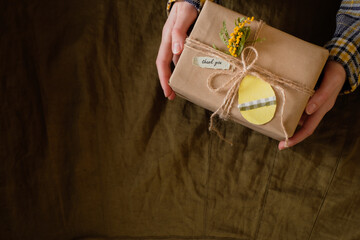 Women's hands holding gift box in craft paper wrapping, with a tag in the shape of an Easter egg, on a dark background. Zero waste and eco friendly concept