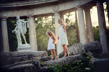 Beautiful blonde long-haired mother and daughter walking like two ancient goddess in the ruins of Colonnade Of Apollo in Pavlovsk park, Russia. Image with selective focus and toning