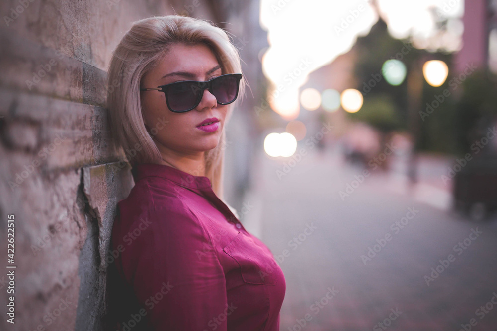 Poster side view of a young blonde female with sunglasses and a pink blouse leaning on a wall