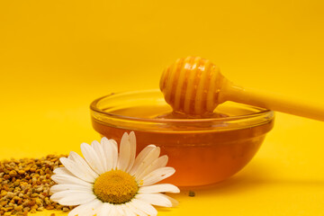 A yellow spoon of honey dipped in a jar of honey and bee bread scattered on a yellow background decorated with live chamomile. Honey dripping around, nice and inviting photos. Healthy food concept.