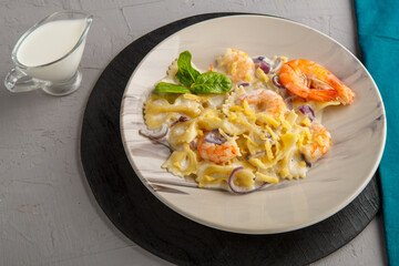 Pasta with shrimps in a creamy sauce on a gray plate on a concrete background next to a blue napkin and a jug of cream.