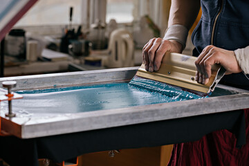 selective focus photo of male hands with squeegee. serigraphy production. printing images on t-shirts by silkscreen method in a design studio - obrazy, fototapety, plakaty