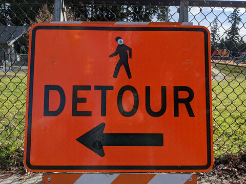 an orange detour sign on a walking path outside a chain link fence