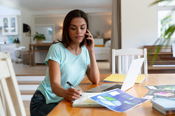 Caucasian woman sitting by desk working from home talking on smartphone and using laptop