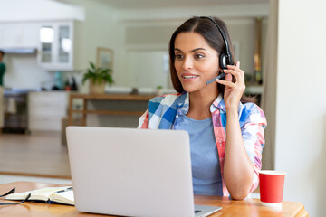 Caucasian woman sitting by desk working from home using laptop and talking on headset