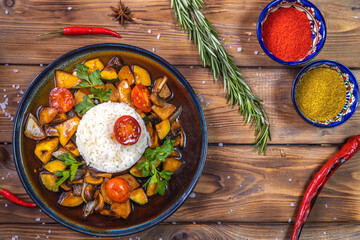 Rice on a plate with stewed vegetables on a wooden background decorated with rosemary, chili pepper, spices.