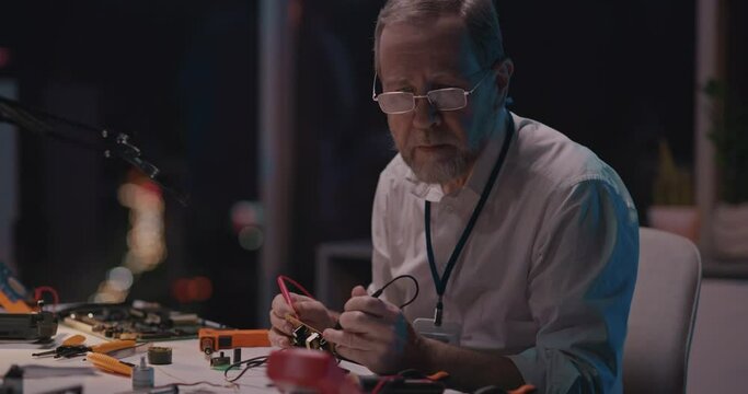 Bearded adult engineer in lab white coat sitting by desk soldering iron circuit board fixing drone device. Technical repair, research office. Engineering.