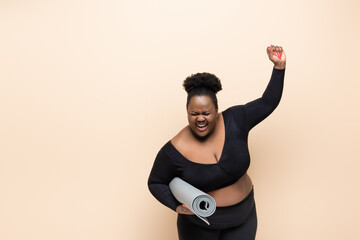 amazed african american plus size woman in sportswear holding fitness mat isolated on beige