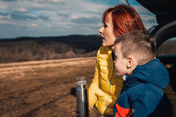 Mother and son take break from driving sitting by the car while drinking tea in nature.