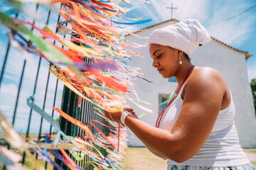 Brazilian woman dressed in the traditional Bahian costume of the Umbanda religion saying a prayer. focus on colored ribbons of religious promises in devotion to Senhor do Bonfim in Bahia, Brazil - obrazy, fototapety, plakaty