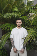 Vertical shot of a tattooed European man in a white t-shirt posing against palm trees