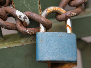 Close up of blue padlock with rusty chain locked at old green gate door, selective focus