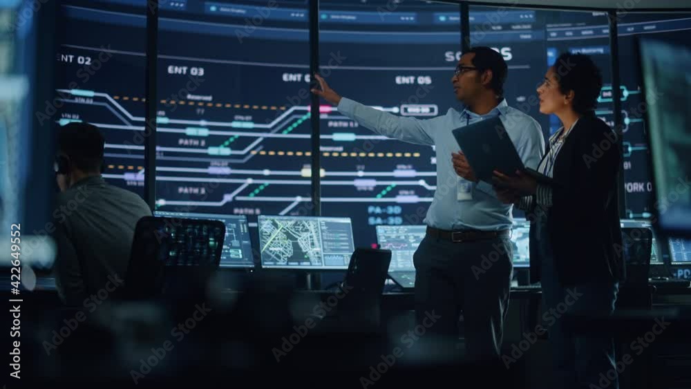 Poster In the System Control Room Project Manager and IT Technical Engineer with Laptop Have Discussion, surrounded by Multiple Monitors with Graphics. Big Monitor Shows Interactive Server Blockchain Info.