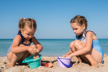 Children girls playing with sand at the beach in summer day. Kids building sand sculptures with shovels and buckets on coast sea. Fun family vacation concept
