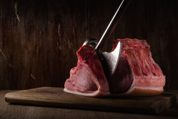 piece of raw pork loin with ribs chopped with an kitchen ax lies on a cutting board with brown old wooden backdrop in shadow. side view. artistic moody photo in rustic style with copy space