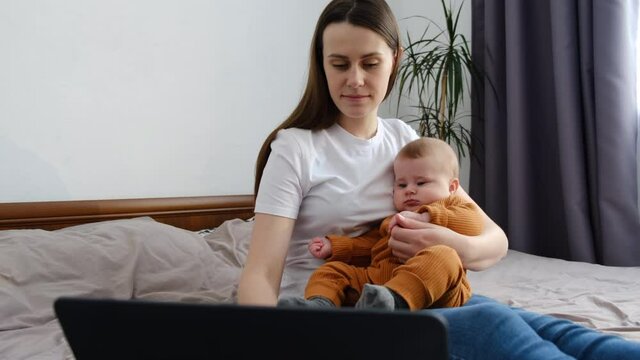 Positive young mother holding small cute baby sitting on bed user laptop, web surfing information on computer, communicating with friends in social network, enjoying free weekend peaceful morning time