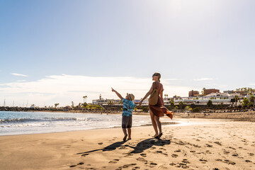 Happy mother and little child holding hands and walking on beach,