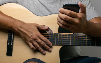 Hands of a dark man on the six strings of a guitar looking at his cell phone looking for new music to play in his home. Wearing a gray t-shirt on a white wall.