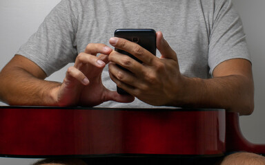 Hands of a dark-haired man on a guitar looking at his cell phone looking for new music to play at his home. Wearing a gray t-shirt on a white wall.