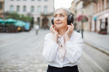 Portrait of happy mature woman standing on city street and listening music in modern wireless headphones. Happy retirement concept.