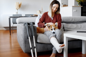 Adult woman in her late twenties on couch at home with crutches and orthopedic plaster caress the dog. Fracture of the leg or foot. Concept of rehabilitation and healing. Orthopedics and Traumatology. - Powered by Adobe