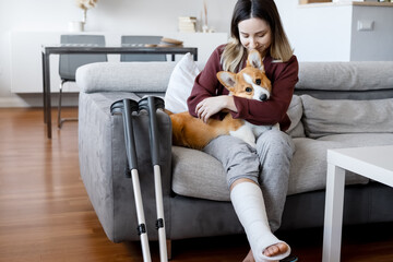 Adult woman in her late twenties on couch at home with crutches and orthopedic plaster caress the...