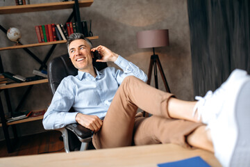Attractive successful gray-haired businessman, freelancer or manager is talking the phone, calling friends or colleagues, throwing his feet on the table, relaxing office, looks away, smiling happily