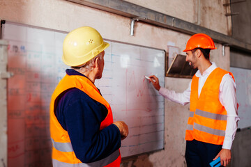 Young engineer and elderly worker in orange vests and hard hat make production plans