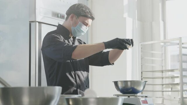 Middle shot portrait of concentrated male confectioner beating eggs for pastry dough in slow motion on sunny kitchen. Professional Caucasian man in Covid-19 face mask working in candy store indoors.